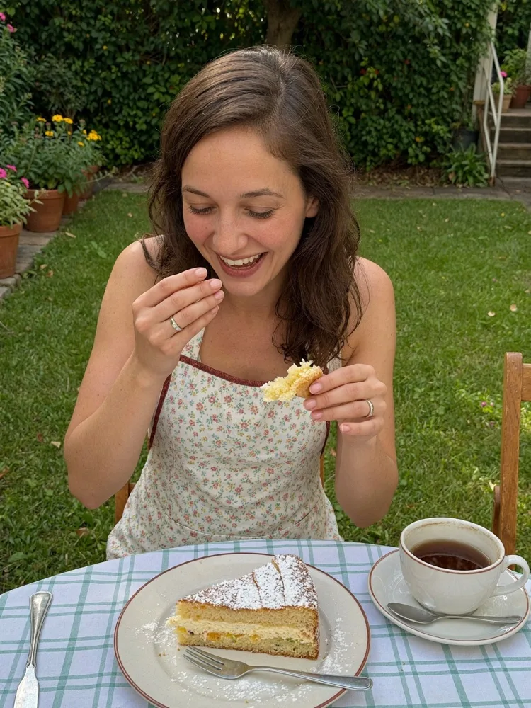 Dieselbe KI-Persona Sophie lachend an einem Gartentisch mit Kuchen und Kaffee.